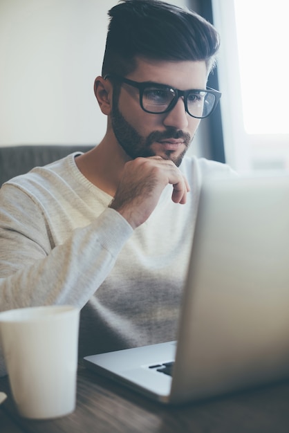Nachdenken über Lösung. Nachdenklicher junger Mann mit Brille, der am Laptop arbeitet und die Hand am Kinn hält, während er im Büro oder Café sitzt