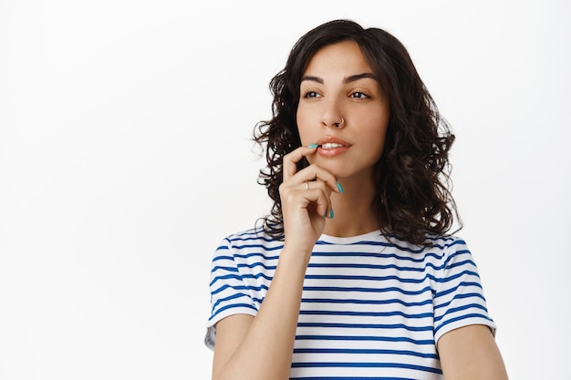 nachdenkendes Mädchen, das beiseite schaut und eine Wahl trifft, die Lippe nachdenklich berühren, nachdenken, auf Weiß in gestreiftem T-Shirt stehen