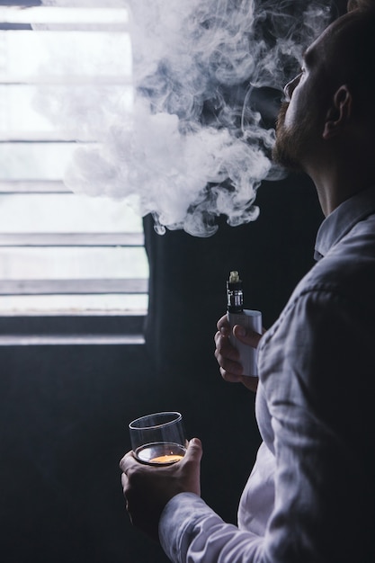 Nachdenklich aussehender Mann in entspannter Stimmung. Mann Freizeit mit Alkohol und Vape im privaten Club, schlechte Angewohnheiten. Luxusleben mit Whisky und Rauchen, dunkler Hintergrund