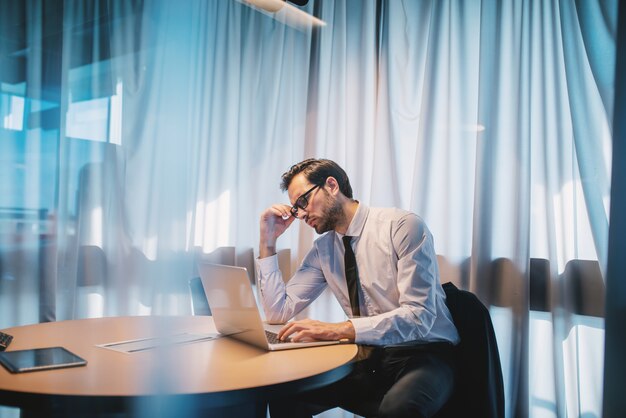 Nachdenklich fokussierter hübscher kaukasischer Geschäftsmann in Hemd und Krawatte und mit Brille, die in seinem modernen Büro sitzen und Berichte lesen. Unternehmensgeschäftskonzept.