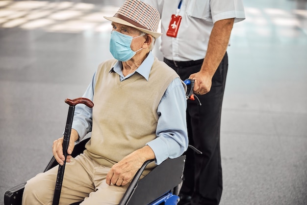 Nachdenklich gealterter kaukasischer männlicher Passagier in einer Einweg-Schutzmaske, der in einem Transportstuhl sitzt