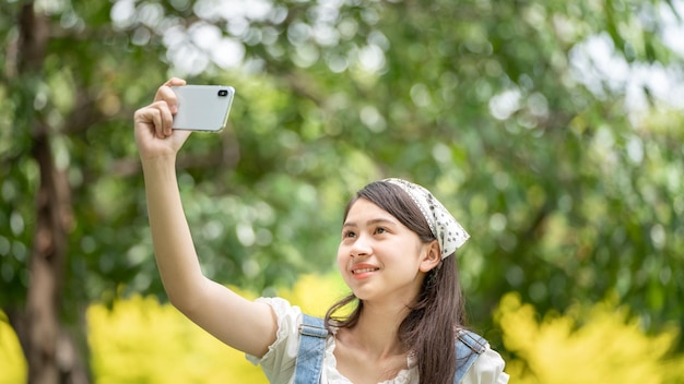 Nachdenklich lächelnde Frau im Park mit Smartphone für Selfie-Foto Porträt eines jungen Charmeurs