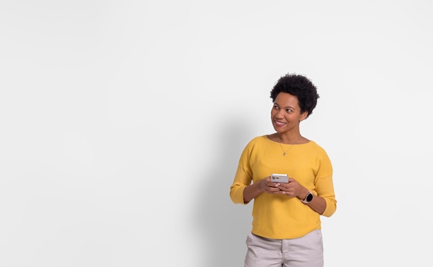 Nachdenklich lächelnde Frau mit Afro-Haaren, die ihr Mobiltelefon in der Hand hält und auf weißem Hintergrund wegschaut