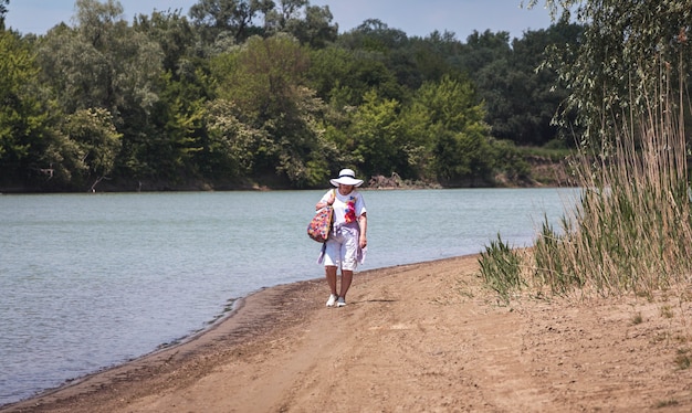 Nachdenkliche ältere Frau, ältere Rentnerin, Ruhe auf dem Sand am Fluss, das Konzept von Zeit und Alter, Sommerferien an der frischen Luft, gesunder Lebensstil