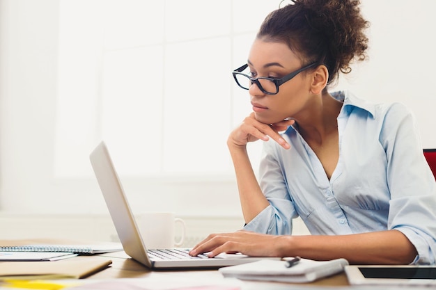 Nachdenkliche afroamerikanische Geschäftsfrau, die im Büro am Laptop arbeitet. Geschäftsfrau, die etwas auf dem Computer tippt, an ihrem Arbeitsplatz sitzt, Platz kopieren