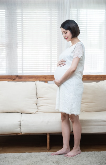 Nachdenkliche asiatische schwangere Frau, die vom Kind träumt, das nahe Fenster steht. Junge glückliche werdende, die an ihr Baby denkt und zukünftiges Leben genießt. Mutterschaft, Schwangerschaft, Glückskonzept, Kopierraum