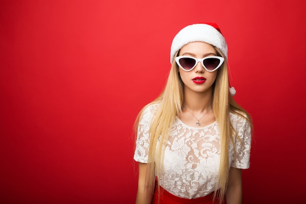 Nachdenkliche Blondine in Weihnachtsmütze auf rotem, isoliertem Hintergrund. Sonnenbrille mit weißem Rand.