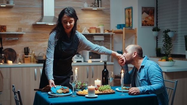 Nachdenkliche Frau, die begeisterten Ehemann dient. Begeisterte Frau, die ein festliches Abendessen mit gesundem Essen zubereitet, für seinen Mann romantisches Abendessen kocht, redet, am Tisch in der Küche sitzt, das Essen genießt