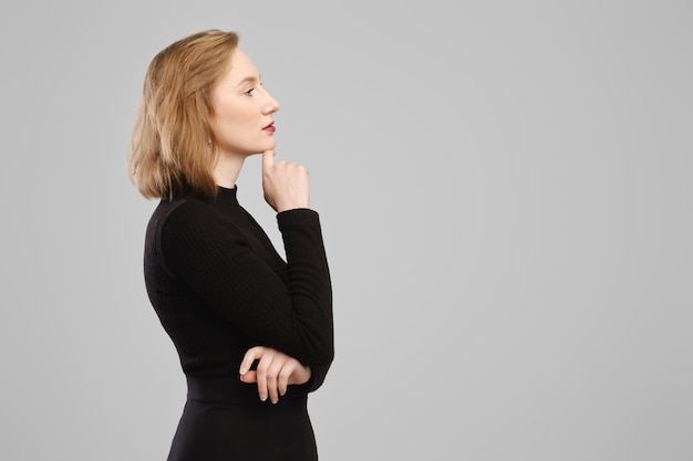Nachdenkliche Frau, die im Profil im Studio über einer grauen Wand steht, das Kinn stützt und nachdenkt
