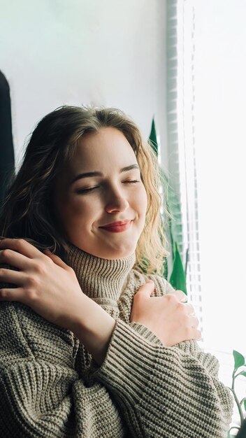 Nachdenkliche Frau, die neben dem Fenster steht und hindurchschaut, sich umarmt und sich einsam fühlt, während sie auf ihren Freund wartet oder von wahrer Liebe und einer gesunden Beziehung träumt