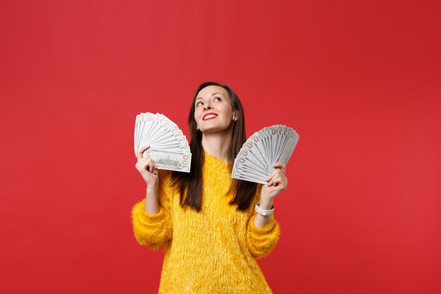 Nachdenkliche Frau in gelbem Pelzpullover, die nach oben schaut, Geldfan in Dollar-Banknoten hält, Bargeld isoliert auf leuchtend rotem Hintergrund. Menschen aufrichtige Emotionen, Lifestyle-Konzept. Kopieren Sie Platz.