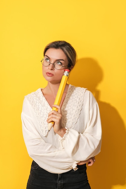 Nachdenkliche Frau mit Brille, die großen Bleistift auf gelbem Hintergrund hält