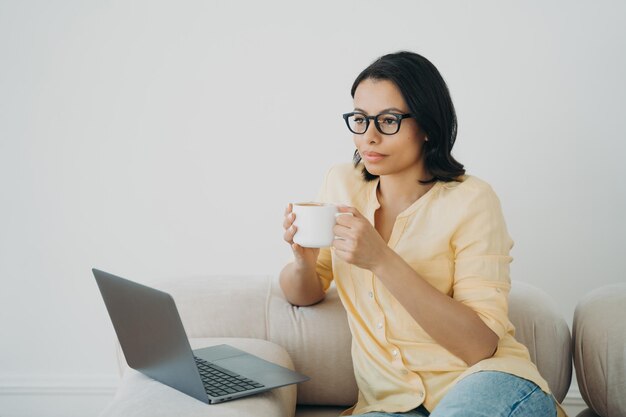 Nachdenkliche Geschäftsfrau arbeitet online auf einem Laptop, der eine Tasse Kaffee im Home Office hält Remote-Job