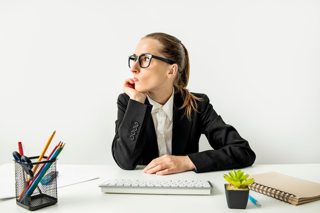 Nachdenkliche Geschäftsfrau in einer Jacke mit Brille und einer Tastatur, die am Arbeitsplatz sitzt