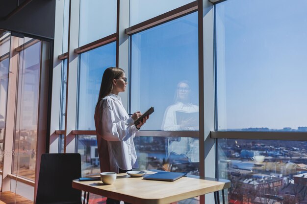 Nachdenkliche Geschäftsfrau mit Notizblock in formeller Kleidung, die in der Nähe eines Panoramafensters in einem Unternehmen steht und über Handelsinvestitionen nachdenkt Nachdenkliche Geschäftsfrau mit Brille