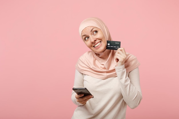 Nachdenkliche junge arabische muslimische Frau in Hijab-leichter Kleidung posiert isoliert auf rosa Hintergrund. Menschen religiösen Islam Lifestyle-Konzept. Mock-up-Kopienbereich. Verwenden Sie das Mobiltelefon und halten Sie eine Kreditkarte bereit.