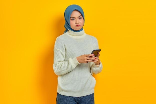 Nachdenkliche junge Asiatin in weißem Pullover und Hijab mit Handy und Blick auf die Kamera isoliert über gelbem Hintergrund Religiöses Konzept des Menschen islam