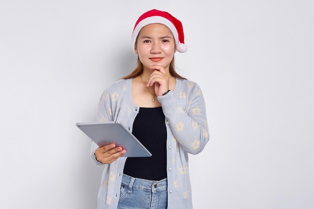 Nachdenkliche junge asiatische Frau in einem Hut Weihnachten mit einem digitalen Tablet und berühren ihre Wange isoliert über weißem Hintergrund Indonesier feiern Weihnachtskonzept