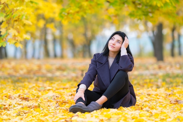 Nachdenkliche junge Frau, die auf dem Boden zwischen Herbstlaub in einem bewaldeten Park sitzt und mit einem ernsten Ausdruck in die Luft starrt