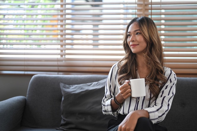 Nachdenkliche junge Frau, die auf dem Sofa sitzt und morgens heißen Kaffee trinkt
