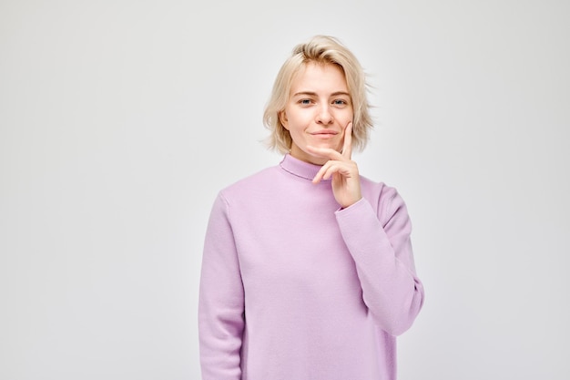 Nachdenkliche junge Frau in einem lila Pullover mit der Hand auf dem Kinn, die auf einem hellen Hintergrund nachdenklich aussieht