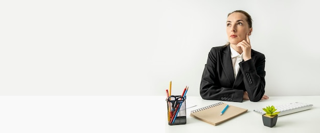 Nachdenkliche junge Frau in Jacke, die am Tisch am Arbeitsplatz sitzt Banner