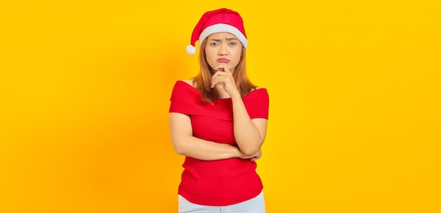 Nachdenkliche junge Frau mit Weihnachtsmütze, die an etwas auf gelbem Hintergrund denkt