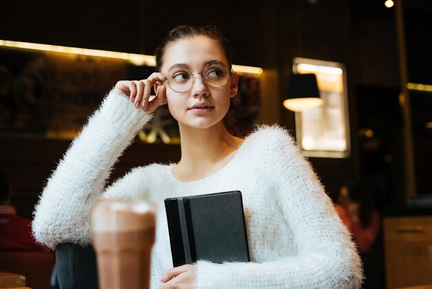 Nachdenkliche junge Freiberuflerin mit Brille, die in einem Café sitzt, ein Notizbuch hält und über die Arbeit nachdenkt