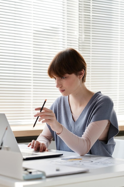 Nachdenkliche junge Geschäftsfrau mit kurzen Haaren, die am Schreibtisch sitzt und Bleistift dreht, während sie mit o arbeitet