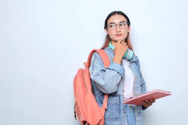 Nachdenkliche junge hübsche Frau, die einen Rucksack trägt, der Bücher hält und über Fragen nachdenkt, die auf weißem Hintergrund isoliert sind