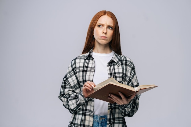 Nachdenkliche junge Studentin, die Seiten des Buches auf grau isoliertem Hintergrund umblättert Hübsches rothaariges Damenmodell, das lässige Kleidung trägt und emotional Gesichtsausdrücke im Studio-Kopienraum zeigt