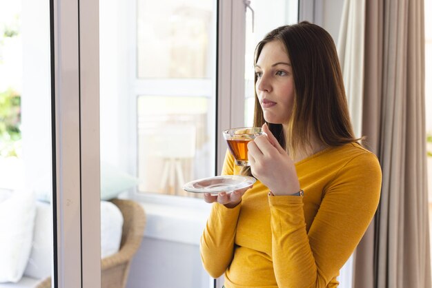 Nachdenkliche kaukasische Frau hält eine Teetasse am Fenster im Wohnzimmer zu Hause