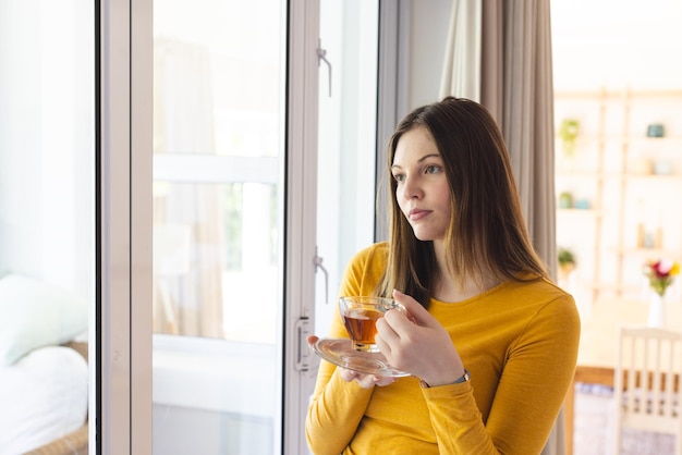 Nachdenkliche kaukasische Frau hält eine Teetasse am Fenster im Wohnzimmer zu Hause