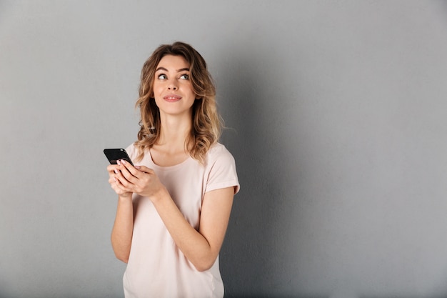 Nachdenkliche lächelnde Frau im T-Shirt, das Smartphone hält und über grauen Boden schaut