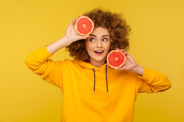 Nachdenkliche, positive, helle Frau mit flauschiger, lockiger Frisur, die eine halbe Scheibe Grapefruit mit nachdenklichem Ausdruck hält, nachdenkt und gesunde, rohe, frische Vitaminnahrung auswählt. Innen, gelber Hintergrund