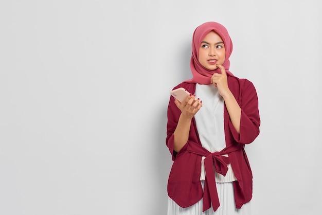 Nachdenkliche schöne asiatische Frau im Freizeithemd mit Handy schaut ernsthaft über etwas nach, das über weißem Hintergrund isoliert ist