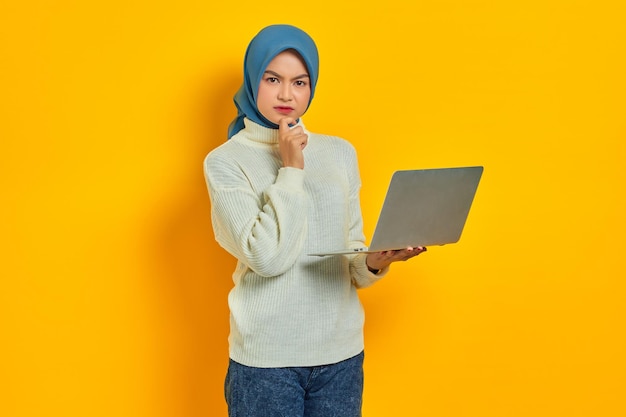 Nachdenkliche schöne asiatische Frau in weißem Pullover mit Laptop sieht ernsthaft über etwas nach, das über gelbem Hintergrund isoliert ist Menschen religiöses Lebensstilkonzept