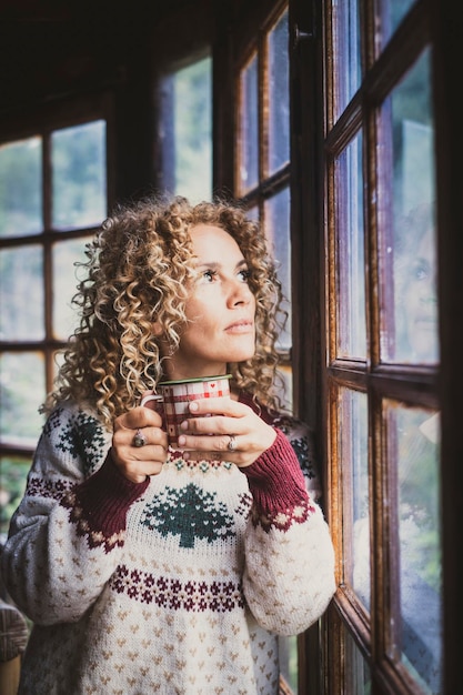 Nachdenkliche süße Frau allein zu Hause, die aus dem Fenster schaut und allein eine Tasse Tee oder Kaffee trinkt Weihnachtsferienzeit Junge weibliche Leute, die Weihnachtspullover tragen und den Vorabend der Dezembersaison genießen