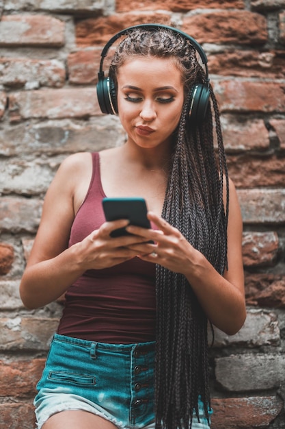 Nachdenkliche süße junge kaukasische Frau mit langen afrogeflochtenen Haaren, die Musik von ihrem Smartphone gegen eine Ziegelwand hört.