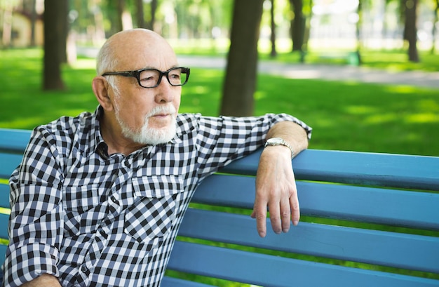 Nachdenklicher älterer Mann im Freien. Älterer Mann in Freizeitkleidung, Brillenträger, sitzend auf der Bank im sonnigen Park, Kopierraum