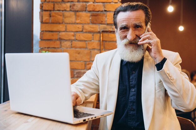 Nachdenklicher älterer Mann mit weißem Bart lächelt, schaut auf seinen Laptop und nimmt telefonisch entgegen. Eine ältere kaukasische Person. Ältere Person und neue Technologie