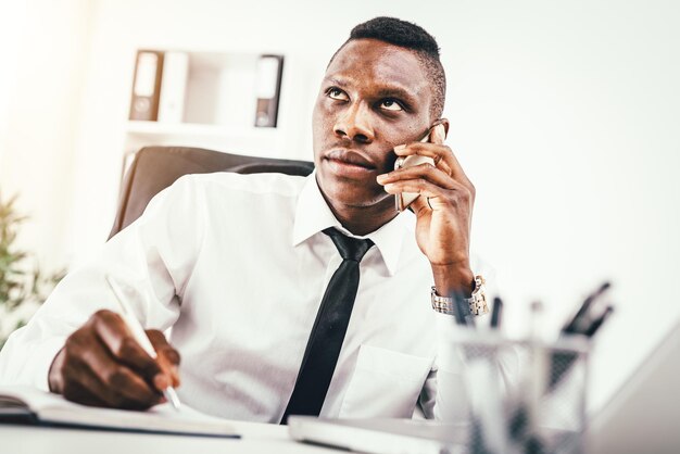 Nachdenklicher afrikanischer Geschäftsmann spricht auf Smartphone und schreibt im Notizbuch im modernen Büro.