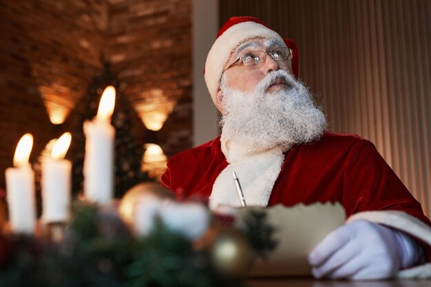 Nachdenklicher alter Weihnachtsmann mit Brille, der am Tisch sitzt und beim Schreiben eines Briefes aufschaut