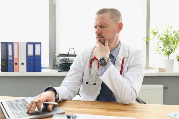 Nachdenklicher Arzt, der in der Klinik auf den Laptop-Bildschirm schaut und auf der Tastatur tippt. Medizinisches Online-Trainingskonzept