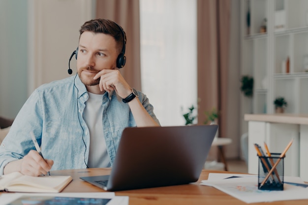 Nachdenklicher bärtiger Freiberufler, der Stift hält und Notizen im Notizbuch macht, während er zu Hause online arbeitet, Kerl in Freizeitkleidung, der an seinem Arbeitsplatz sitzt und über die Arbeit nachdenkt. Remote-Job-Konzept