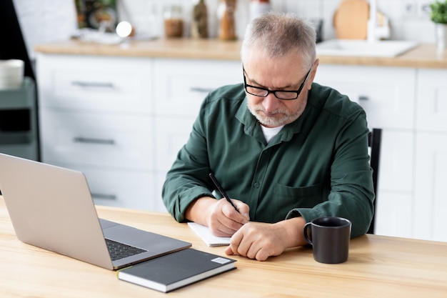 Foto nachdenklicher ernster älterer mann mit laptop, der am tisch sitzt, überprüft die finanzen oder lernt online zu hause