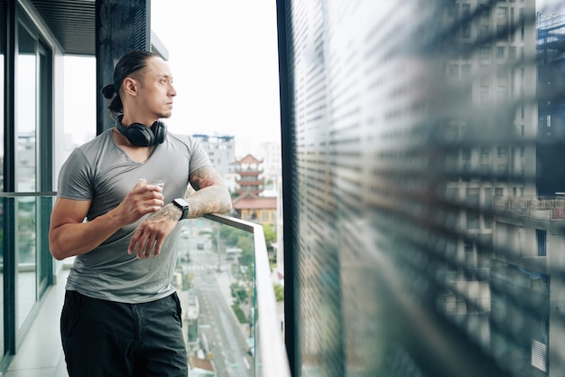 Nachdenklicher ernsthafter fitter Mann, der mit einem Glas Whisky auf dem Balkon der Wohnung steht und den Blick auf die Stadt genießt