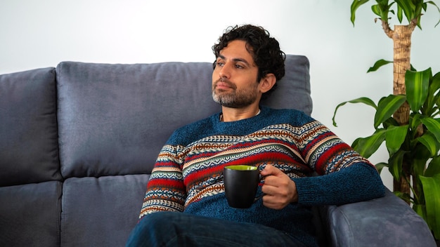 Nachdenklicher erwachsener Mann, der morgendlichen Expresso-Kaffee trinkt, sich entspannt und die Freizeit genießt, während er auf der Couch zu Hause sitzt. Ruhiger, sorgloser bärtiger Mann, der ein Café hat und auf dem Sofa sitzt.