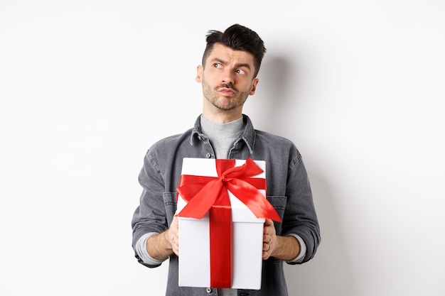 Nachdenklicher Freund schaut beiseite und hält Geschenkbox, wartet auf Liebhaber, macht Überraschungsgeschenk am Valentinstag, Planungsdatum mit Freundin, weißer Hintergrund.