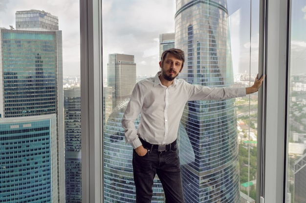 Nachdenklicher Geschäftsmann mit Bart steht neben dem Fenster im Büro vor dem Hintergrund der Wolkenkratzer in Moskau in weißem Hemd und Weste